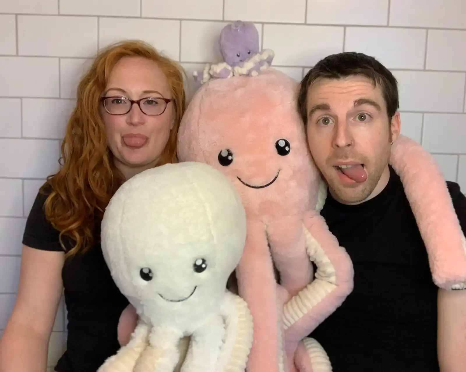 Photograph of a redheaded woman with glasses wearing a black t-shirt (Sarah) and a short-haired man (Matt), both sticking out their tongues non-seriously, surrounded by stuffed octopus toys of various sizes.